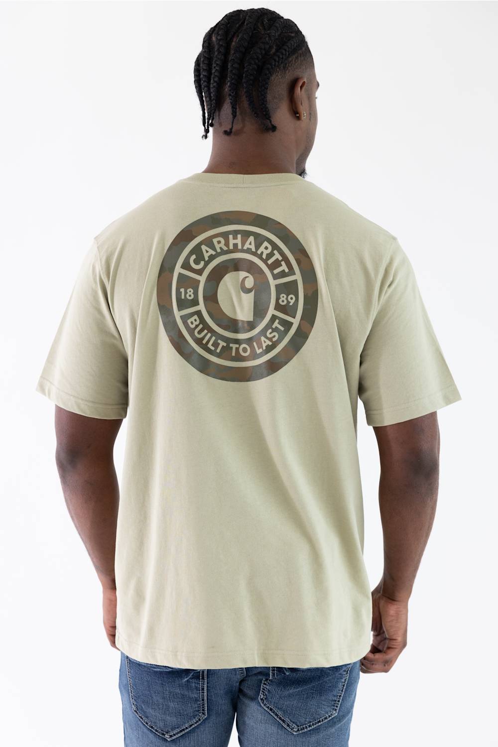A man viewed from behind wearing a light beige Carhartt t-shirt with a large circular logo on the back, paired with blue jeans against a white studio background.