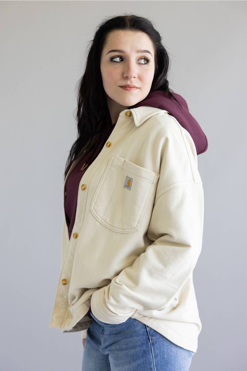 Woman wearing a beige Carhartt jacket over a maroon hoodie with a plain background