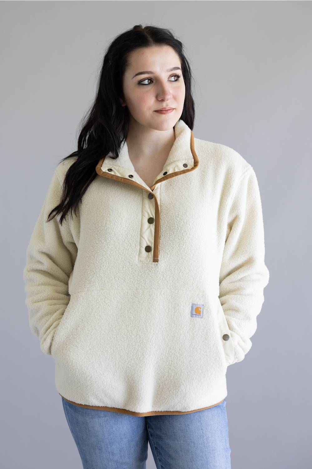 A woman wearing a cream-colored fleece pullover sweater with brown trim and buttons, paired with blue jeans, standing against a solid gray background.