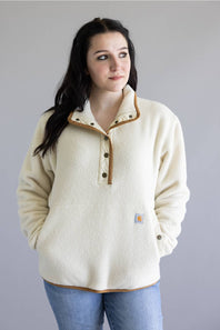 A woman wearing a cream-colored fleece pullover sweater with brown trim and buttons, paired with blue jeans, standing against a solid gray background.