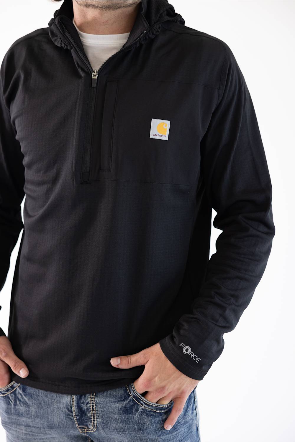 A male model standing against a white background, wearing a black Carhartt Force 1/2 zip hooded sweatshirt over a white t-shirt and blue jeans.