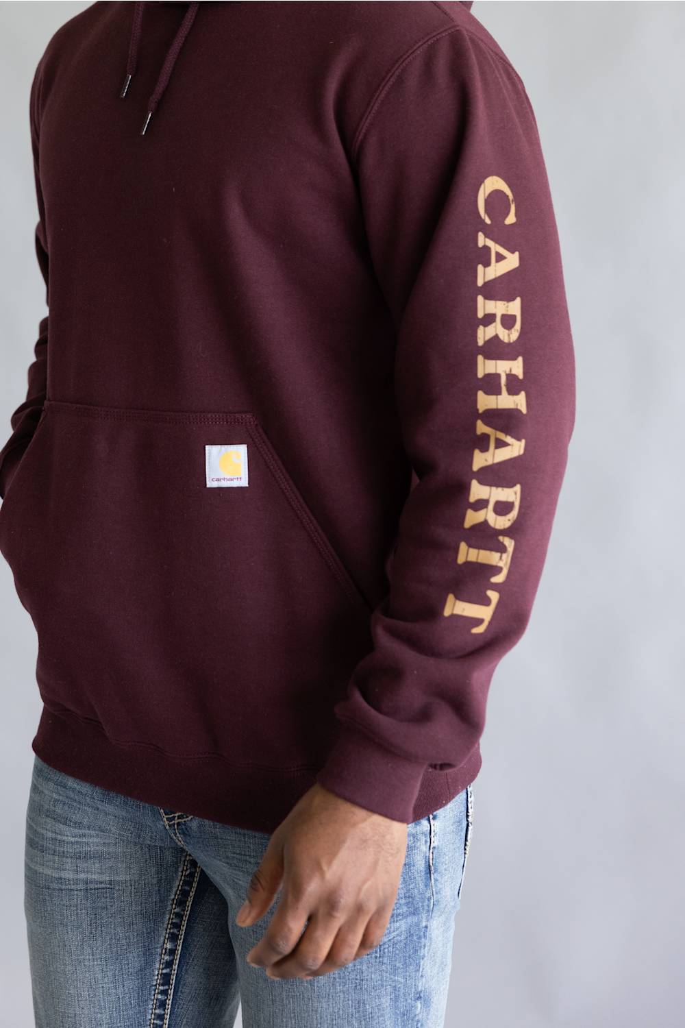 A person wearing a maroon Carhartt hoodie with gold 'CARHART' text printed down the right sleeve and a small Carhartt logo patch on the front pocket, paired with blue jeans against a light gray background.