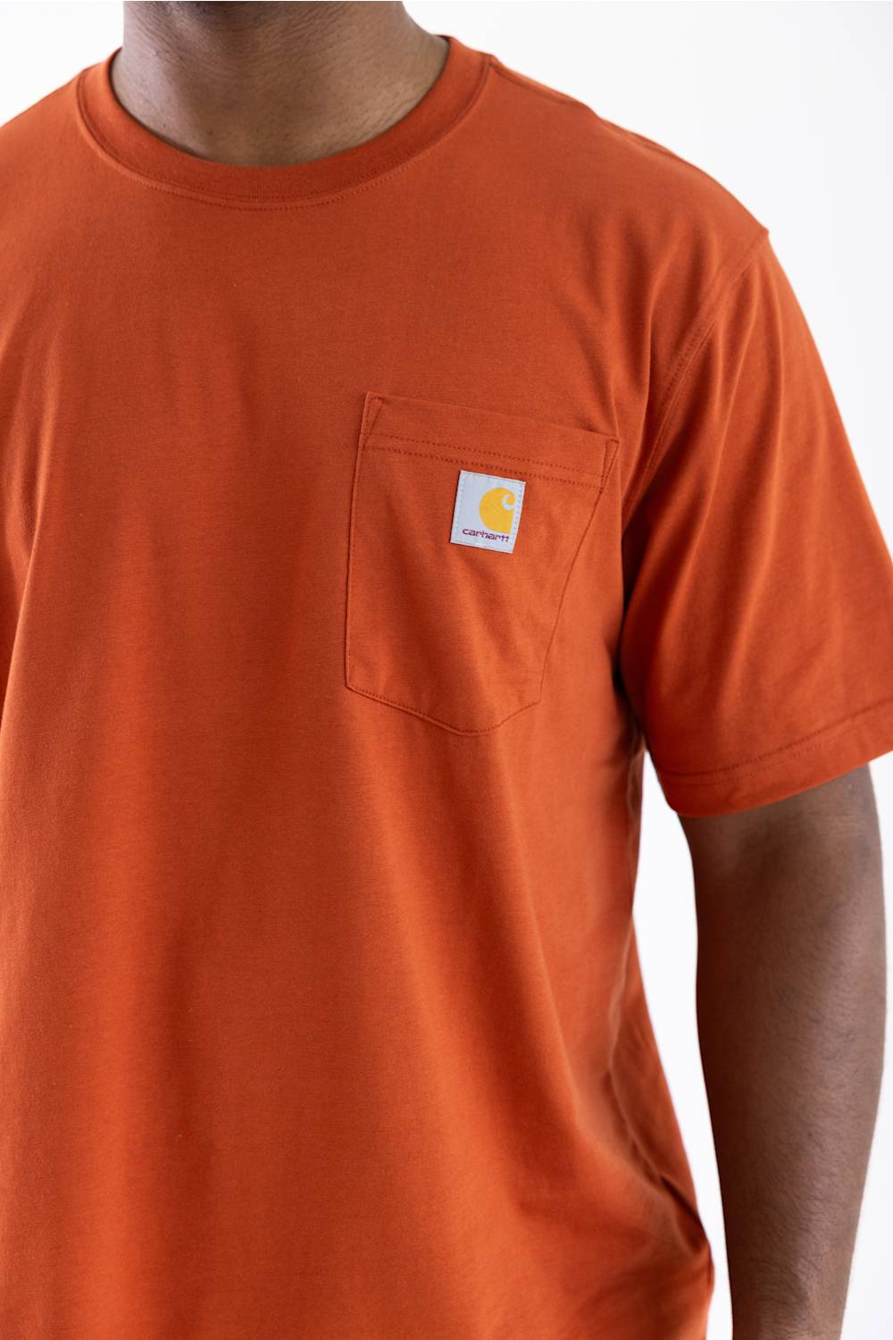 A close-up of a person wearing a bright orange short-sleeve crewneck pocket t-shirt. The shirt features a classic Carhartt logo patch on the left chest pocket, set against a clean white background.