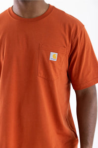 A close-up of a person wearing a bright orange short-sleeve crewneck pocket t-shirt. The shirt features a classic Carhartt logo patch on the left chest pocket, set against a clean white background.
