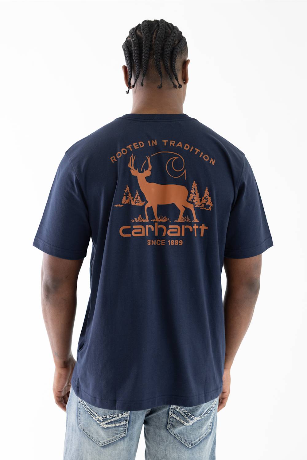 A man viewed from behind wearing a navy blue Carhartt t-shirt with a large orange graphic on the back, paired with light wash jeans against a white studio background.