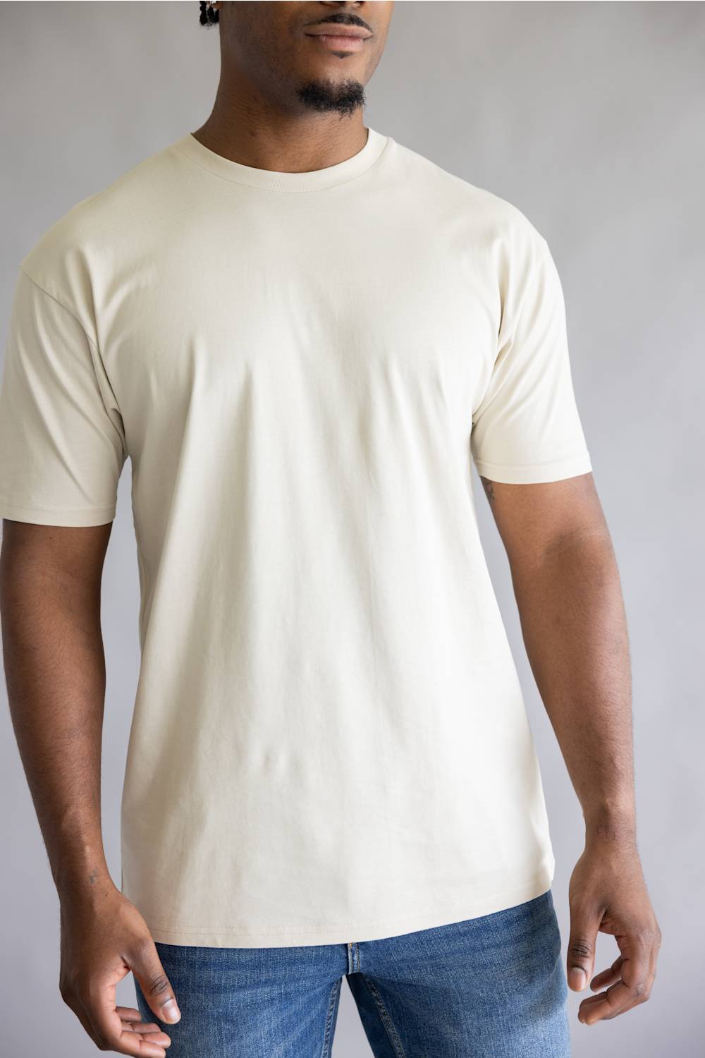 A man wearing a plain cream-colored short-sleeve crewneck t-shirt and blue jeans, standing against a neutral gray studio background.