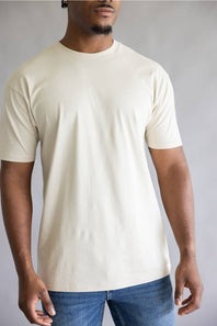 A man wearing a plain cream-colored short-sleeve crewneck t-shirt and blue jeans, standing against a neutral gray studio background.