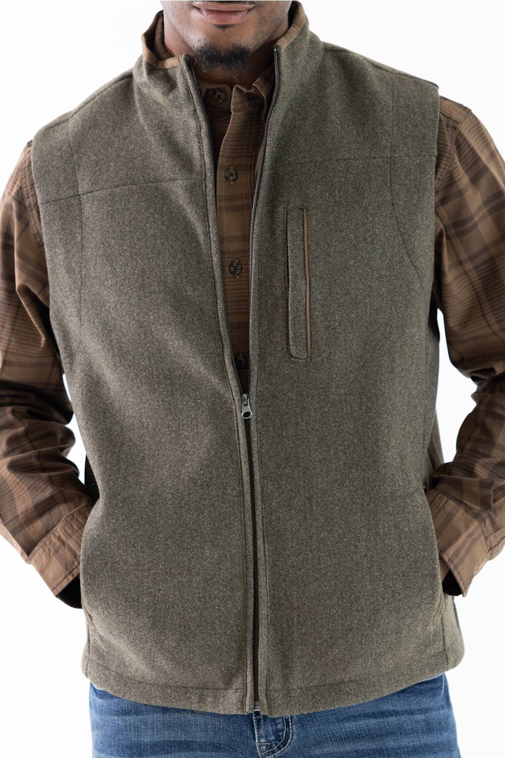 A man wearing an olive green zip-up vest over a brown plaid button-down shirt and blue jeans, shown from the chin down against a white background.