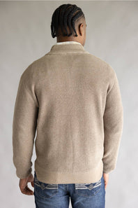 A man viewed from behind wearing a beige ribbed knit sweater over a white collared shirt and blue jeans, set against a plain light gray studio background.