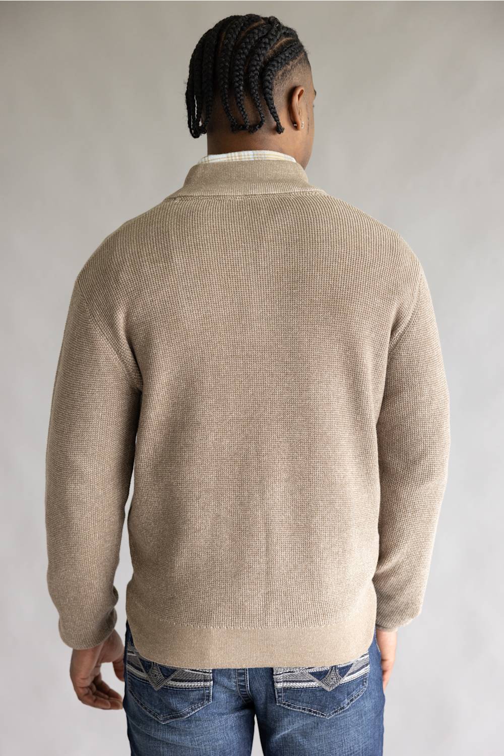 A man viewed from behind wearing a beige ribbed knit sweater over a white collared shirt and blue jeans, set against a plain light gray studio background.