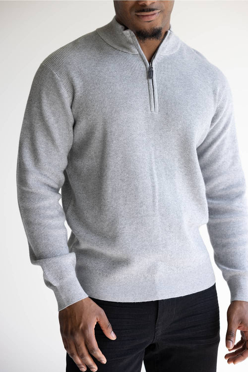 A man wearing a light gray ribbed knit quarter-zip sweater with a stand collar, paired with black pants, shown against a plain light background.