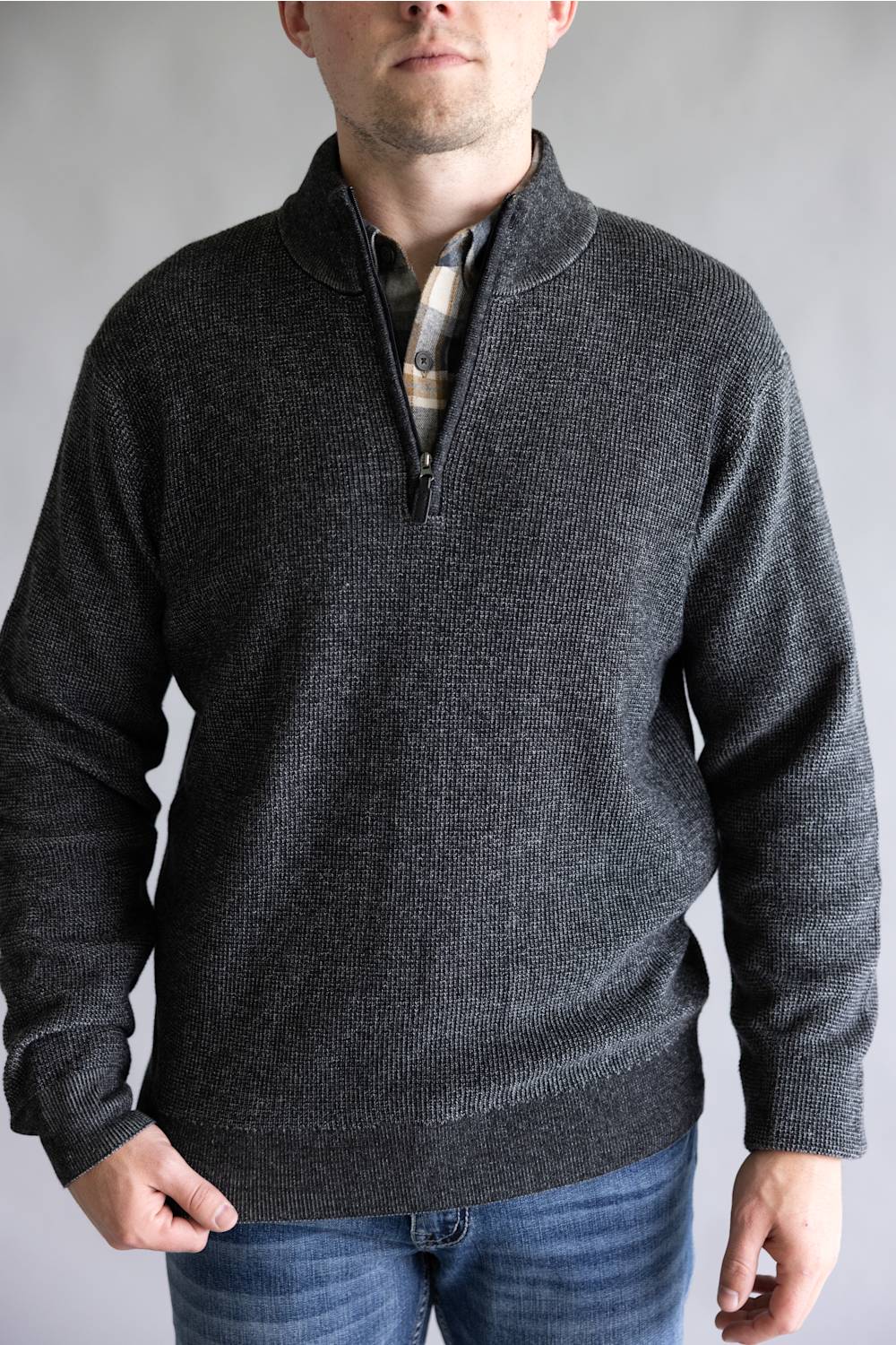 A man wearing a dark charcoal gray ribbed knit quarter-zip sweater over a patterned collared shirt and blue jeans, photographed against a neutral gray studio background.