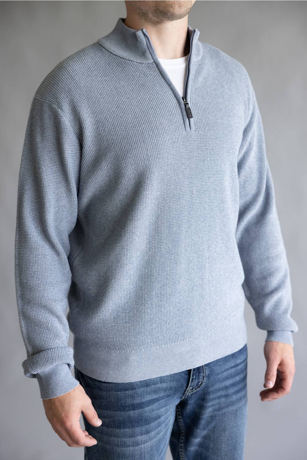 A man wearing a light gray ribbed knit quarter-zip sweater over a white undershirt and blue jeans, shown from the neck down against a neutral gray background.
