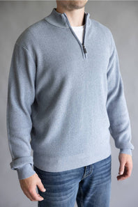 A man wearing a light gray ribbed knit quarter-zip sweater over a white undershirt and blue jeans, shown from the neck down against a neutral gray background.
