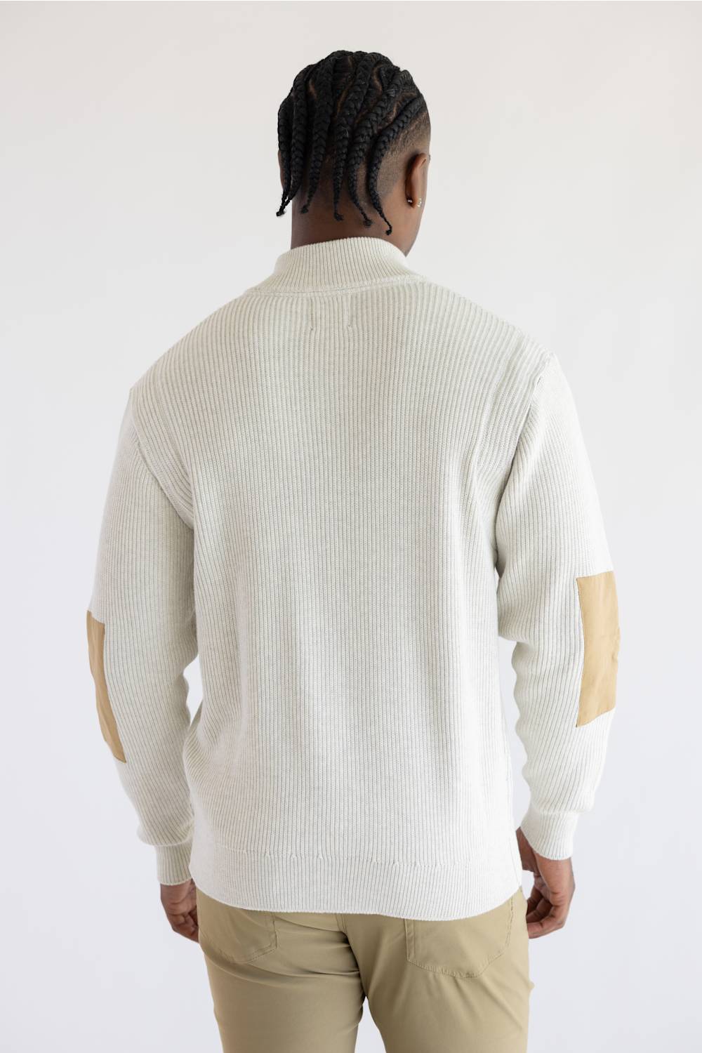 A man viewed from behind wearing a cream ribbed knit sweater with tan elbow patches and khaki pants, set against a plain light gray studio background.