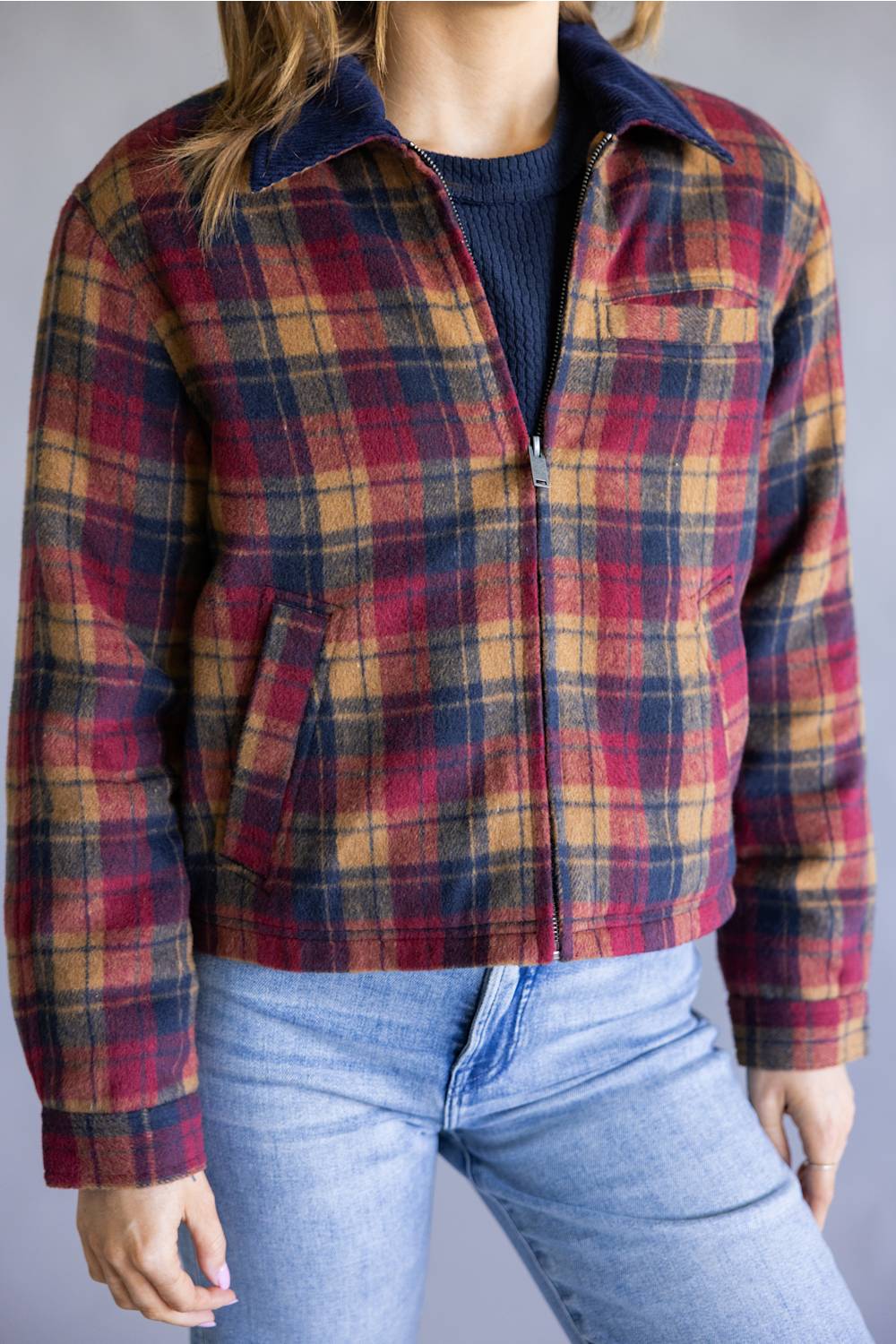 A person wearing a cropped zip-front plaid jacket in red, mustard, and navy tones, paired with a dark ribbed top and light blue jeans, against a plain light gray background.