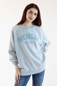 A woman with red hair wearing a light blue washed sweatshirt with 'MICHIGAN GREAT LAKES EST. 1977' printed in blue on the front, paired with blue jeans, against a plain white background.