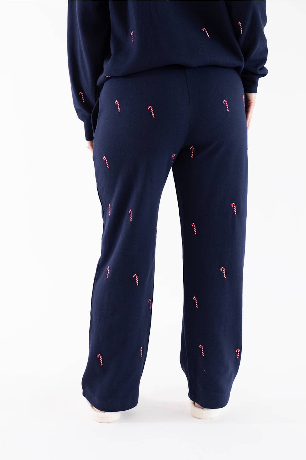 Close-up back view of a woman wearing a navy blue fleece crewneck sweatshirt and matching straight-leg sweatpants, both featuring an all-over embroidered red and white candy cane pattern.