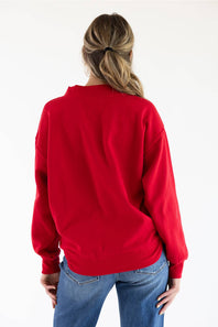 A woman viewed from behind wearing a bright red crewneck sweatshirt and blue jeans against a plain white background.