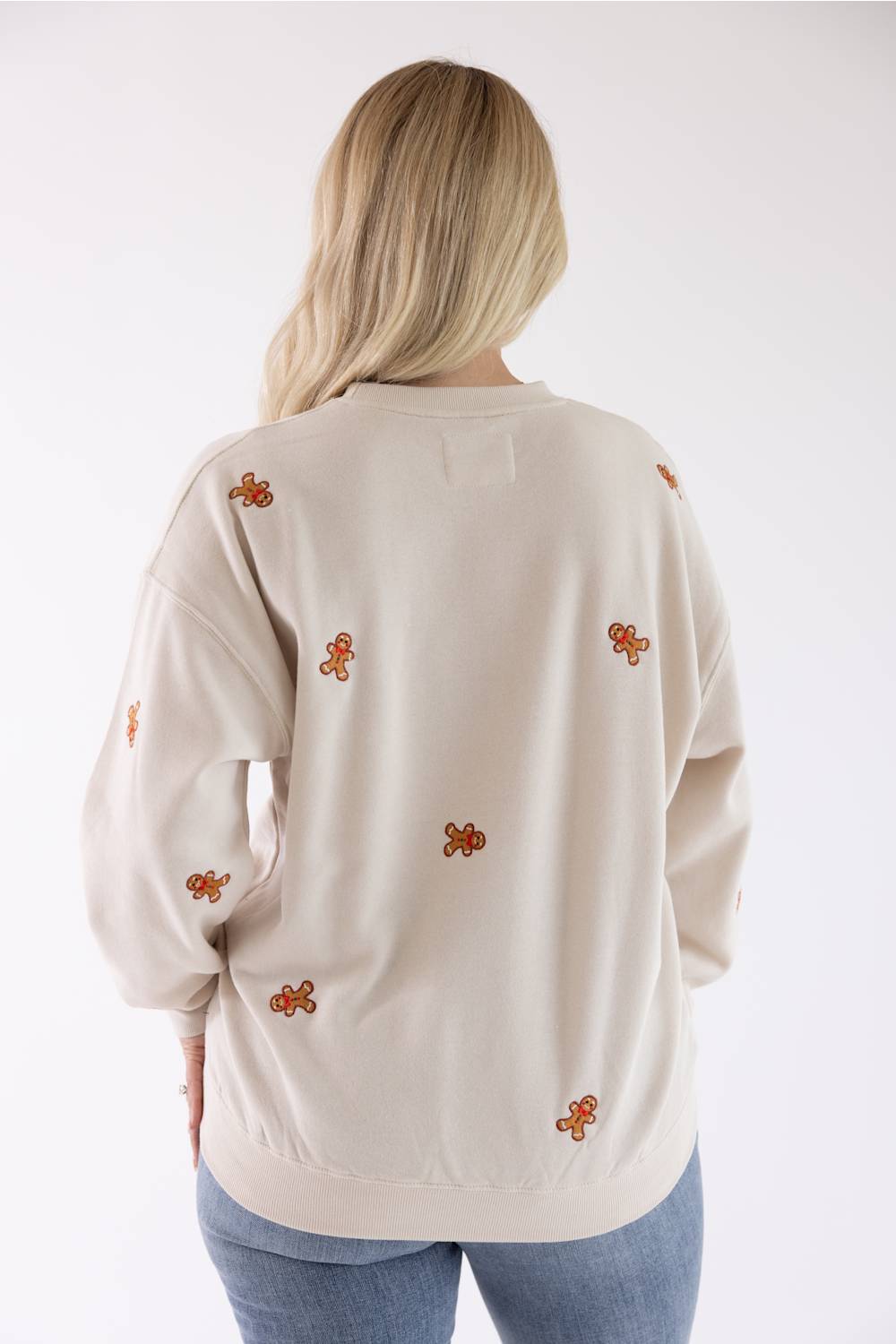 Back view of a woman wearing a beige crewneck fleece sweatshirt with an all-over embroidered gingerbread man pattern.