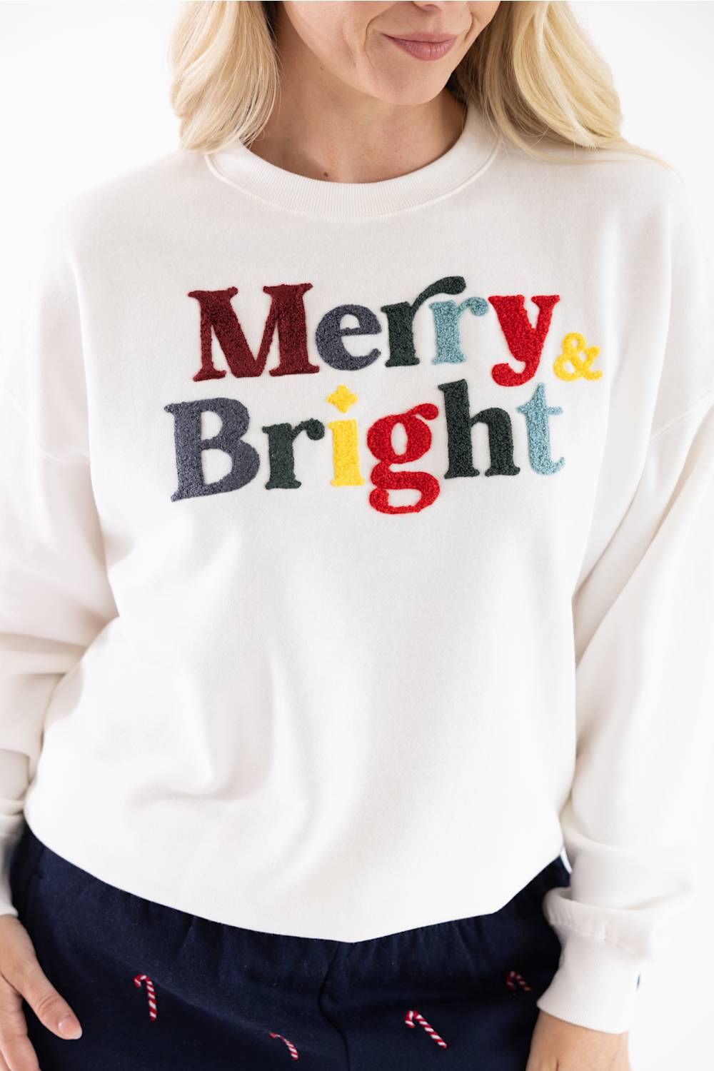 A smiling blonde woman wearing a white crewneck sweatshirt with colorful chenille "Merry & Bright" text across the chest, paired with dark blue lounge pants.