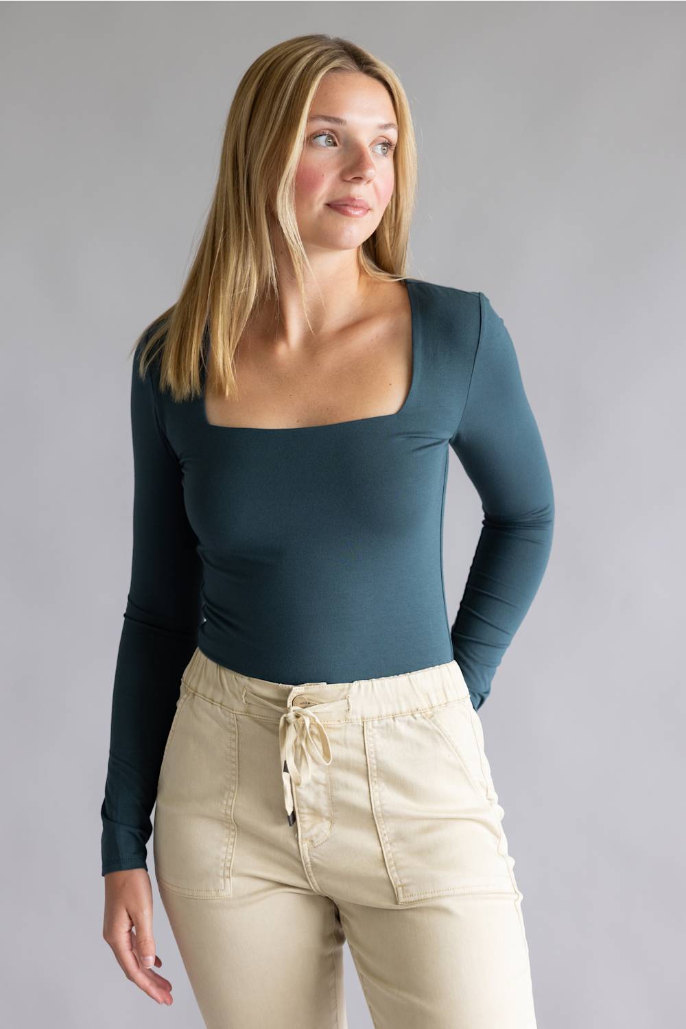 A woman modeling a long-sleeve scoop-neck top in a muted teal blue, paired with beige drawstring pants, against a solid gray studio background.