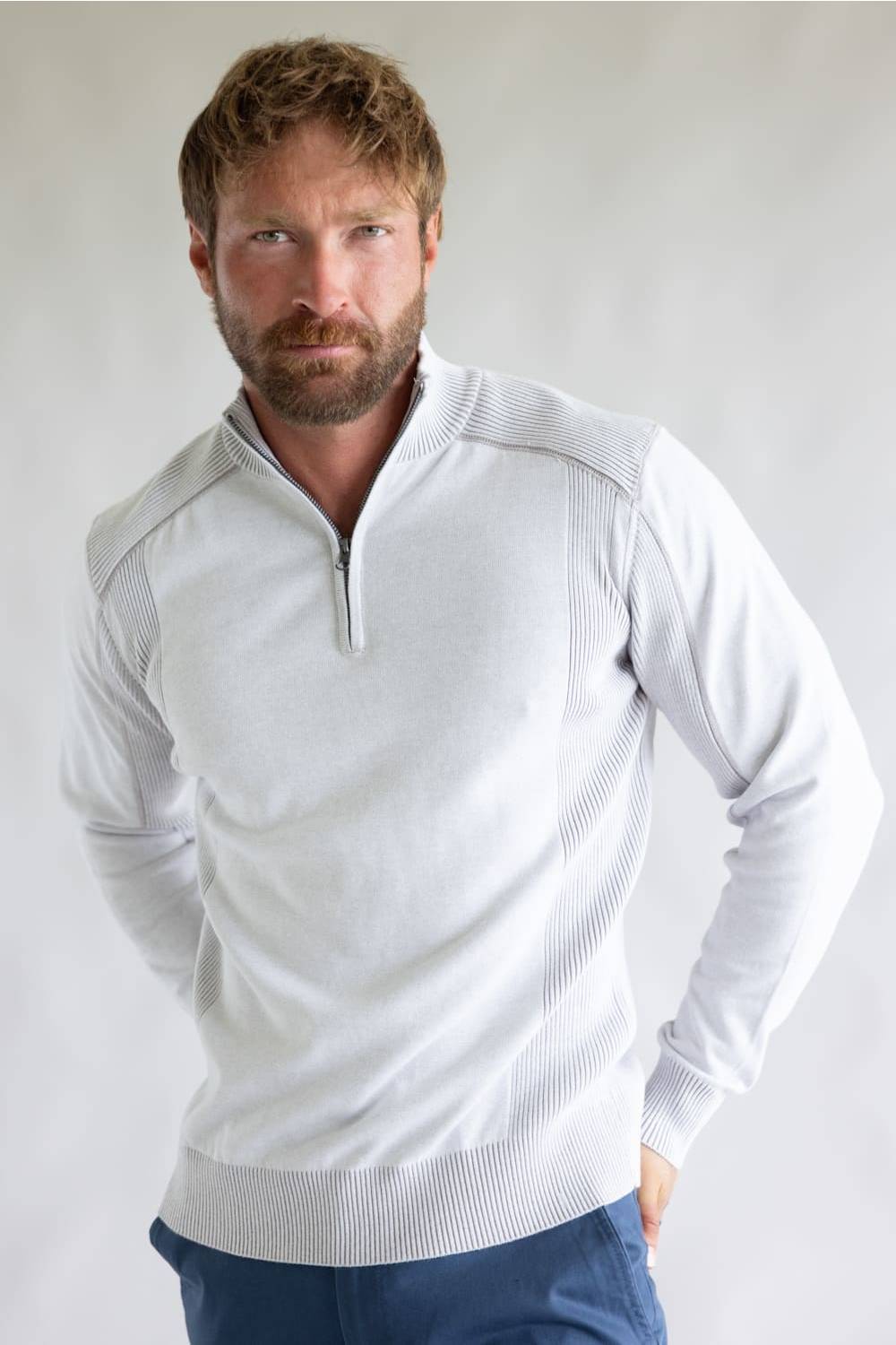 A man wearing a light gray quarter-zip knit sweater with ribbed trim, paired with blue pants, photographed against a neutral light gray studio background.