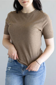 A woman wearing a short-sleeve crewneck knit top in taupe brown, paired with blue jeans, shown from the neck down against a plain light gray background.