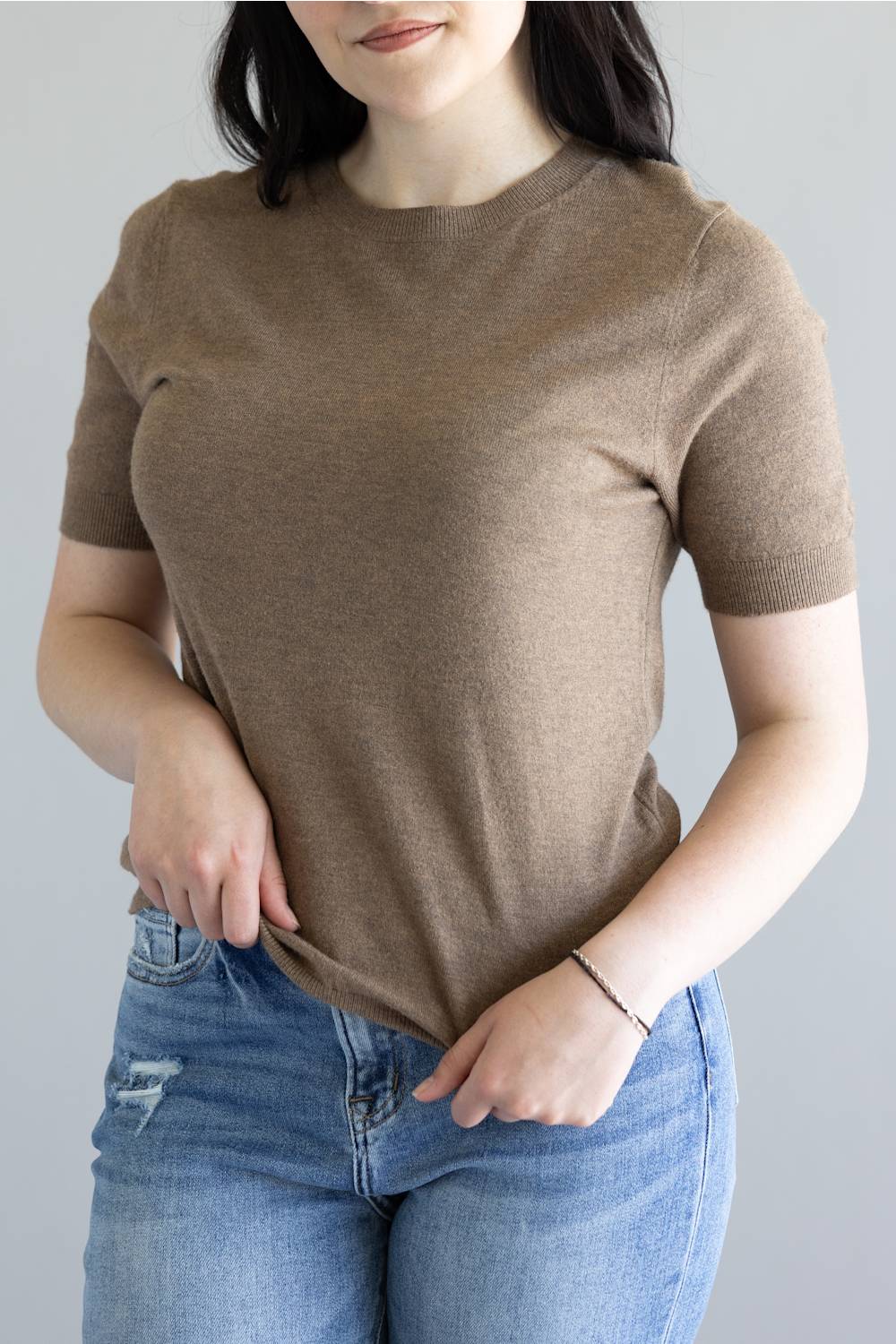 A woman wearing a short-sleeve crewneck knit top in taupe brown, paired with blue jeans, shown from the neck down against a plain light gray background.