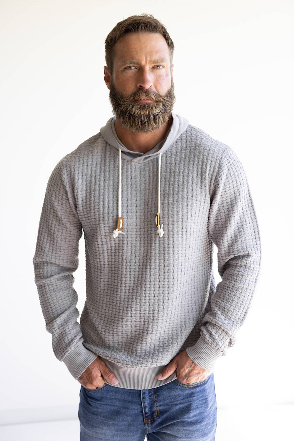 A man with a full beard wearing a gray waffle-knit hoodie with white drawstrings and blue jeans, standing against a plain white background.