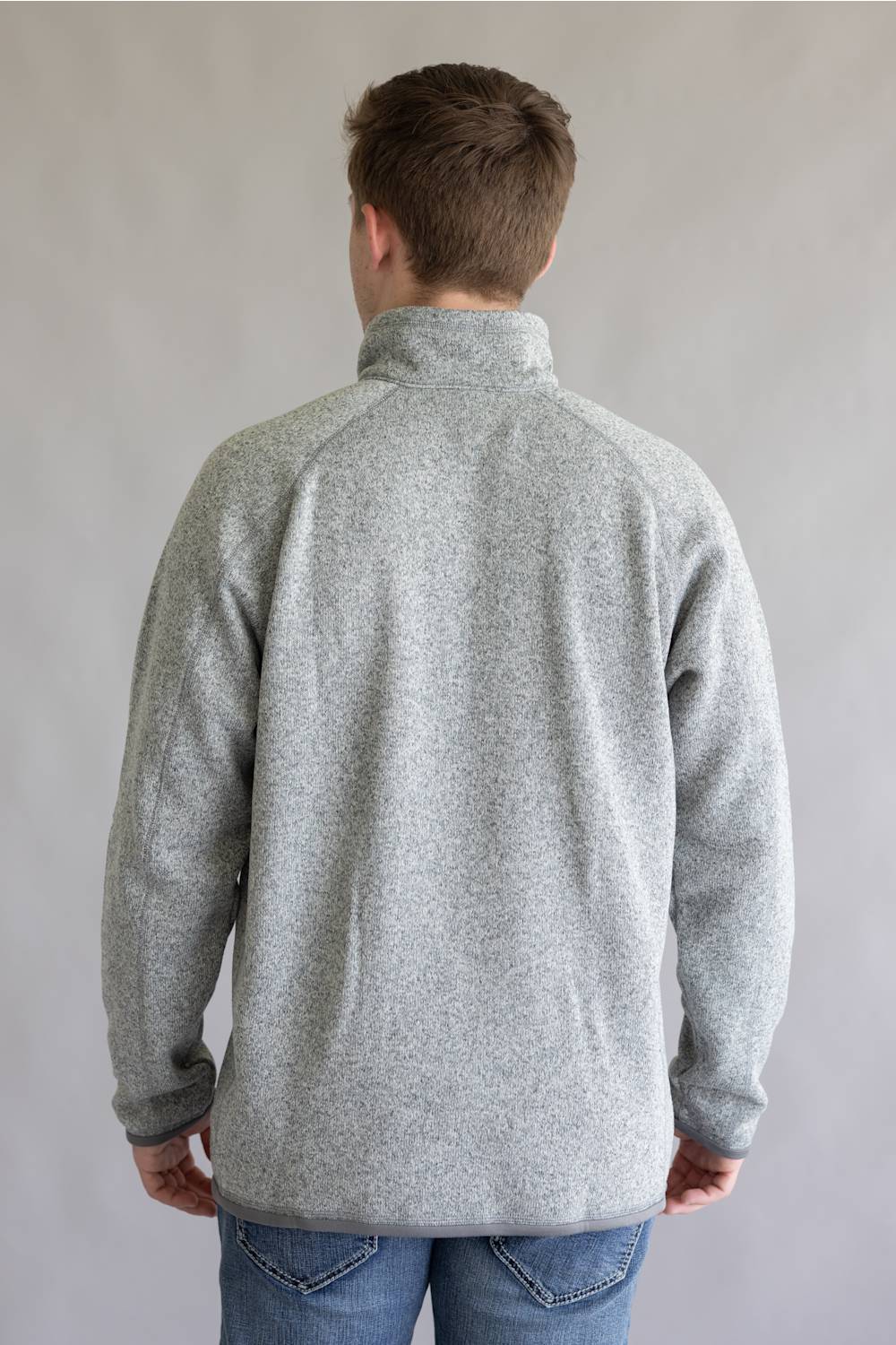 A man viewed from behind wearing a light gray heathered quarter-zip fleece pullover and blue jeans, standing against a neutral gray studio background.
