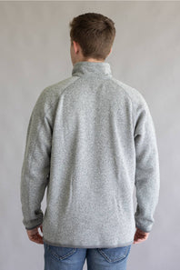A man viewed from behind wearing a light gray heathered quarter-zip fleece pullover and blue jeans, standing against a neutral gray studio background.
