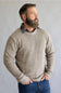 A man with a full beard wearing a textured, waffle-knit beige crewneck sweater over a dark plaid collared shirt and blue jeans, standing against a neutral gray background.