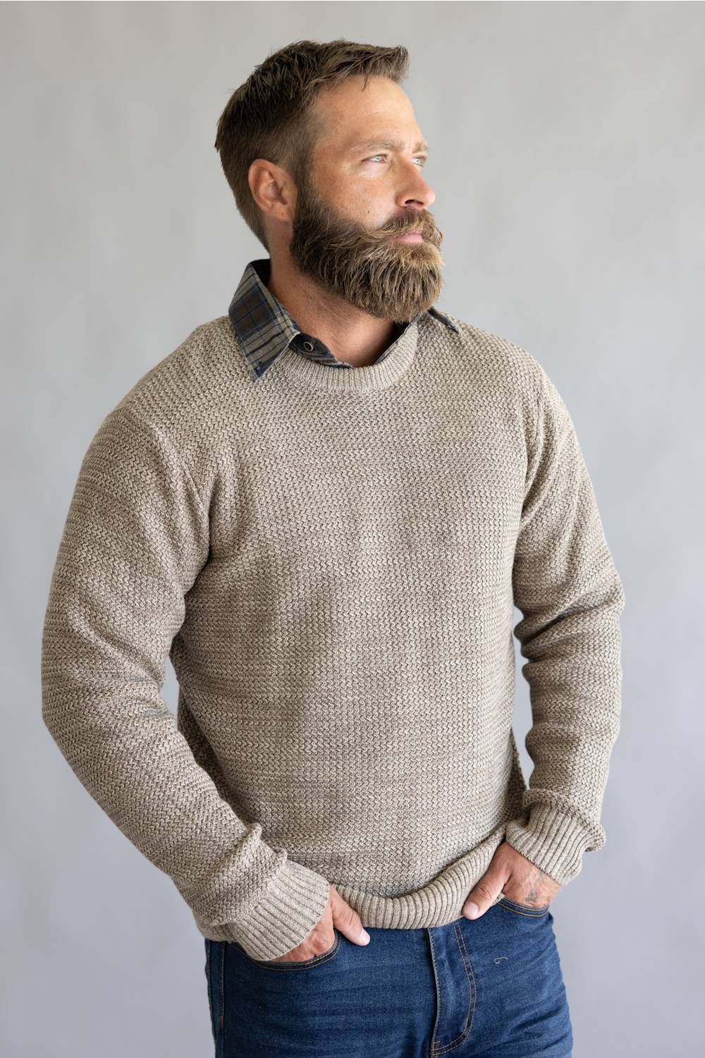 A man with a full beard wearing a textured, waffle-knit beige crewneck sweater over a dark plaid collared shirt and blue jeans, standing against a neutral gray background.