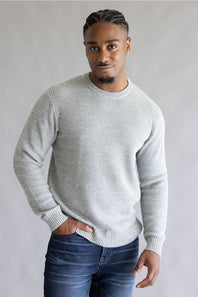 A man wearing a light gray textured knit crewneck sweater and dark blue jeans, standing against a neutral gray background.