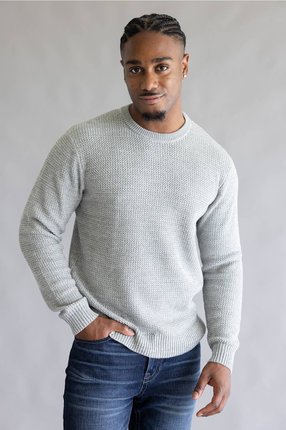 A man wearing a light gray textured knit crewneck sweater and dark blue jeans, standing against a neutral gray background.