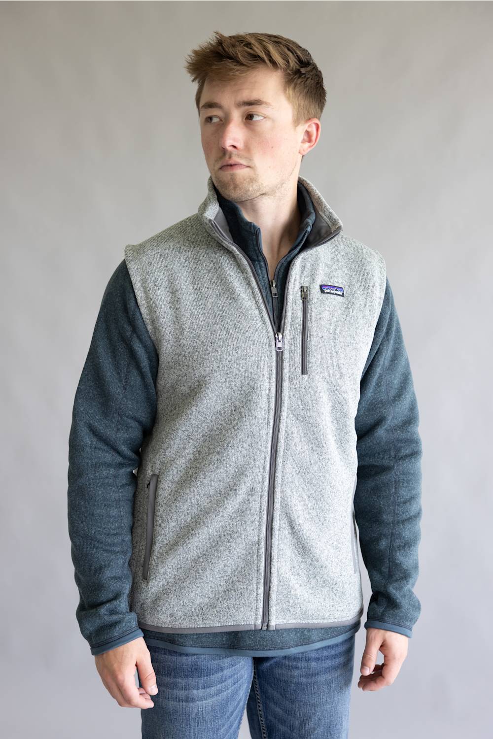 A man wearing a Patagonia fleece vest with a heather gray body and dark blue sleeves, paired with blue jeans, standing against a neutral gray background.