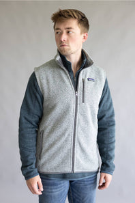 A man wearing a Patagonia fleece vest with a heather gray body and dark blue sleeves, paired with blue jeans, standing against a neutral gray background.