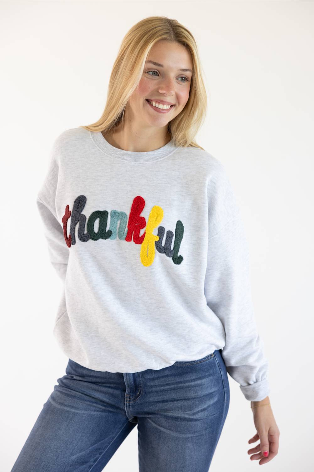 A smiling blonde woman wearing a light gray crewneck sweatshirt with the word 'thankful' written across the chest in red, green, blue, and yellow, paired with blue jeans against a plain white background.