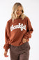 A woman wearing a rust-brown crewneck sweatshirt with 'thankful' printed in white script across the chest, paired with brown and white striped pants, against a plain white background.