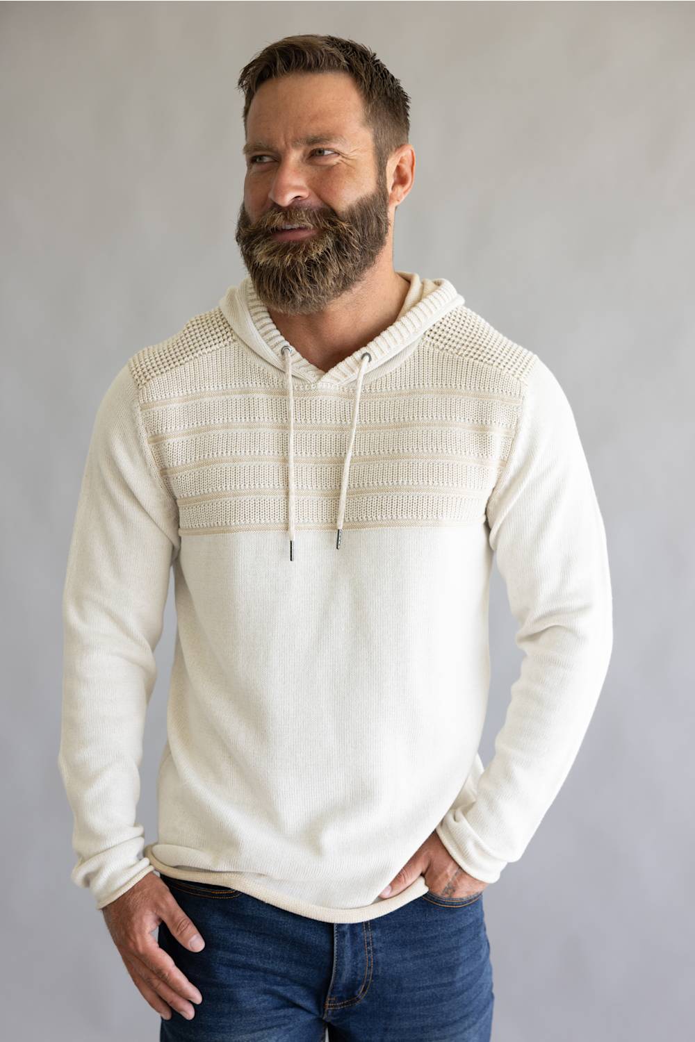 A man wearing a cream-colored knit hoodie with a textured, ribbed yoke across the chest and hood, paired with blue jeans, against a neutral gray background.