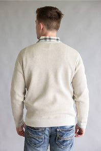 A man viewed from behind wearing a cream-colored ribbed knit sweater over a plaid collared shirt and blue jeans, set against a plain gray studio background.