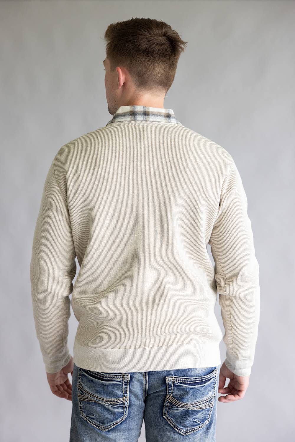 A man viewed from behind wearing a cream-colored ribbed knit sweater over a plaid collared shirt and blue jeans, set against a plain gray studio background.