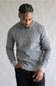 A man wearing a light gray ribbed knit mock neck sweater and black pants, standing against a neutral light gray background.