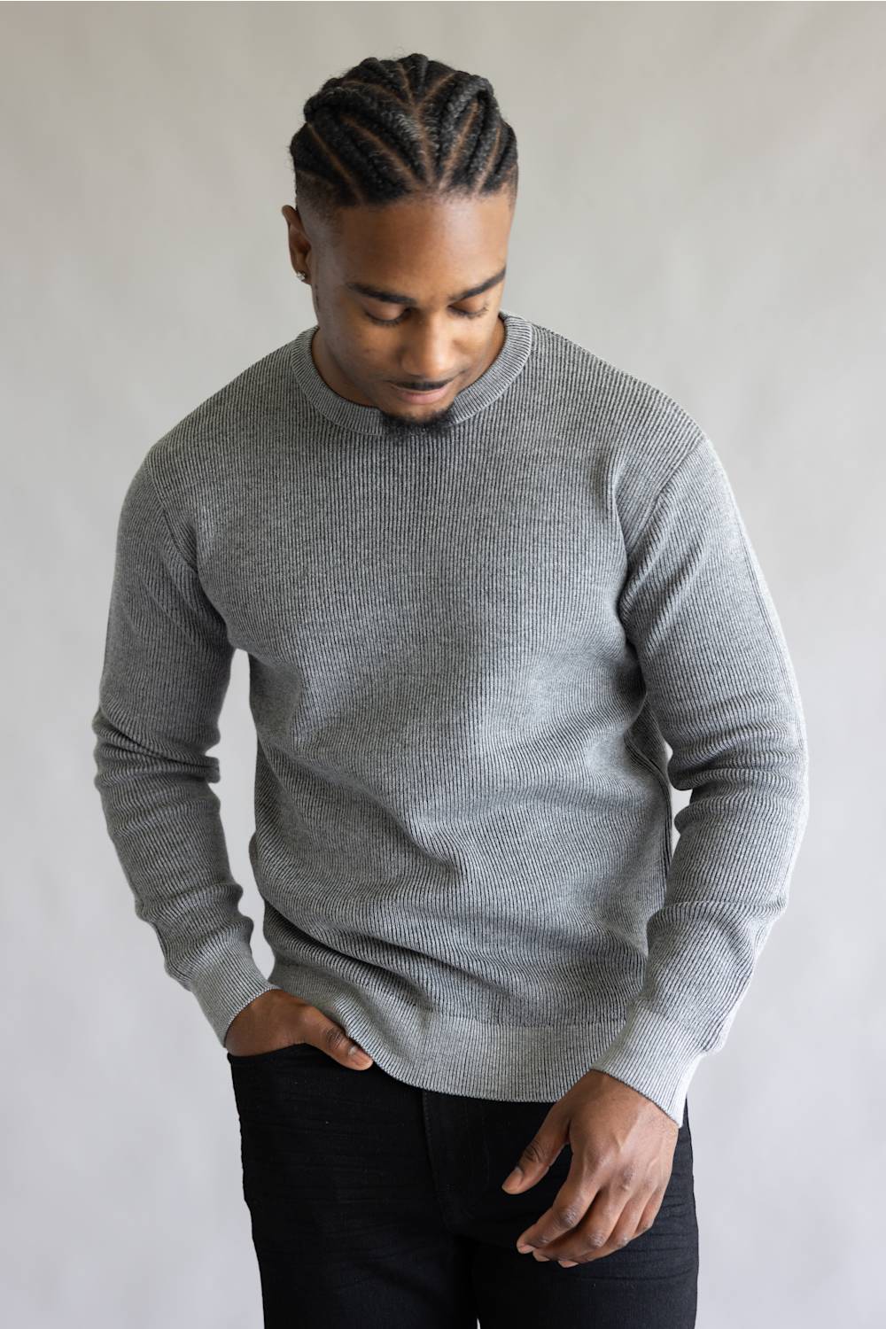 A man wearing a light gray ribbed knit mock neck sweater and black pants, standing against a neutral light gray background.
