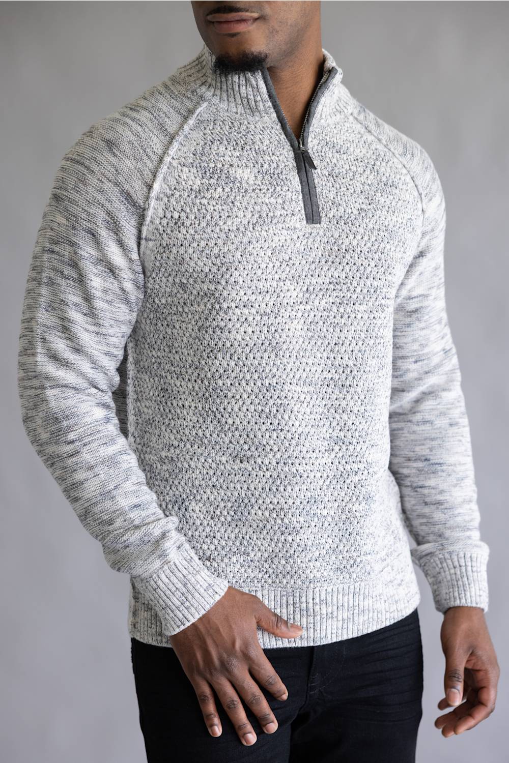 A man wearing a light gray and white heathered knit quarter-zip sweater with a dark gray zipper, paired with black pants, against a neutral gray background.
