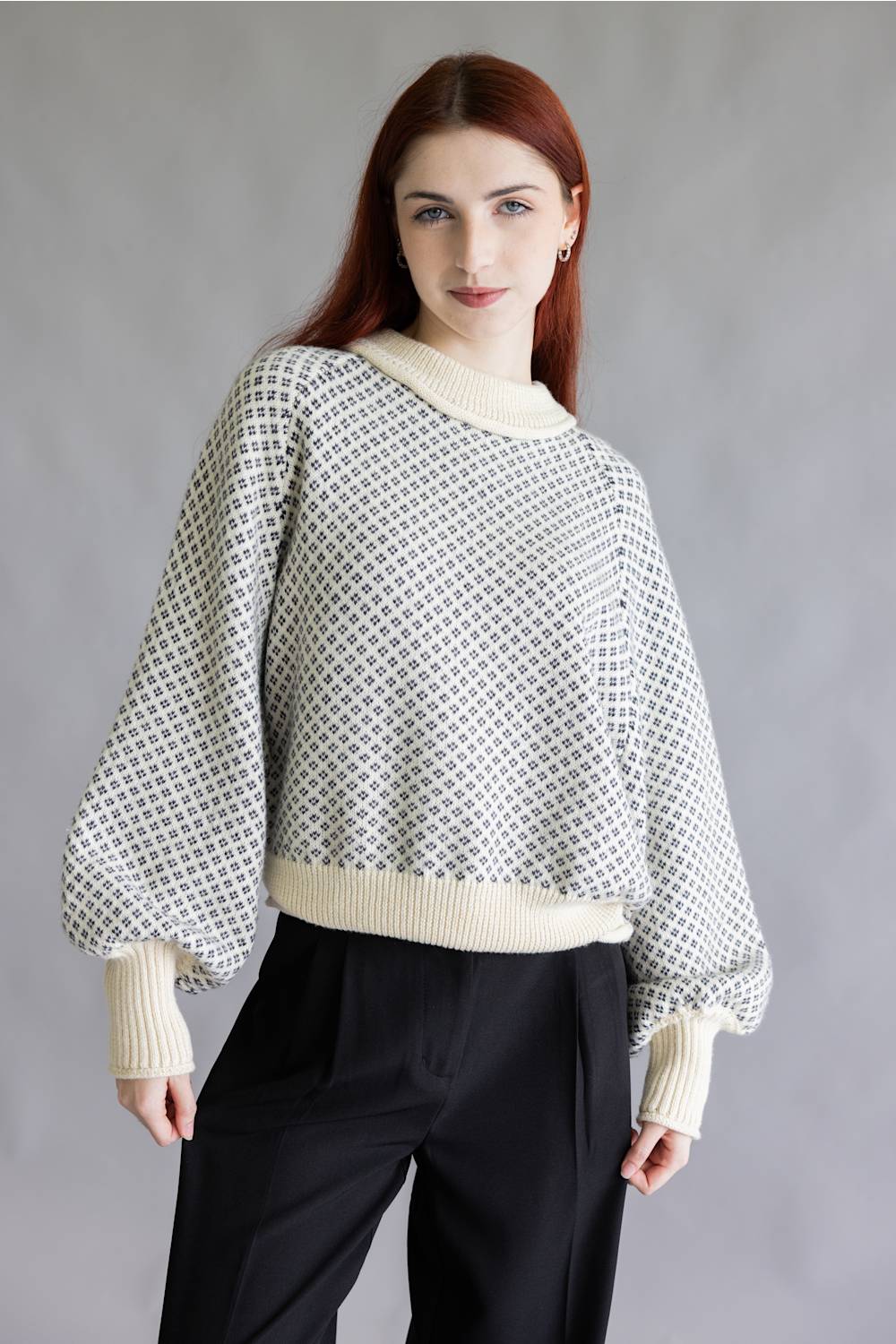 A woman with red hair wearing a cream and black patterned knit sweater with balloon sleeves and black trousers, standing against a gray studio background.