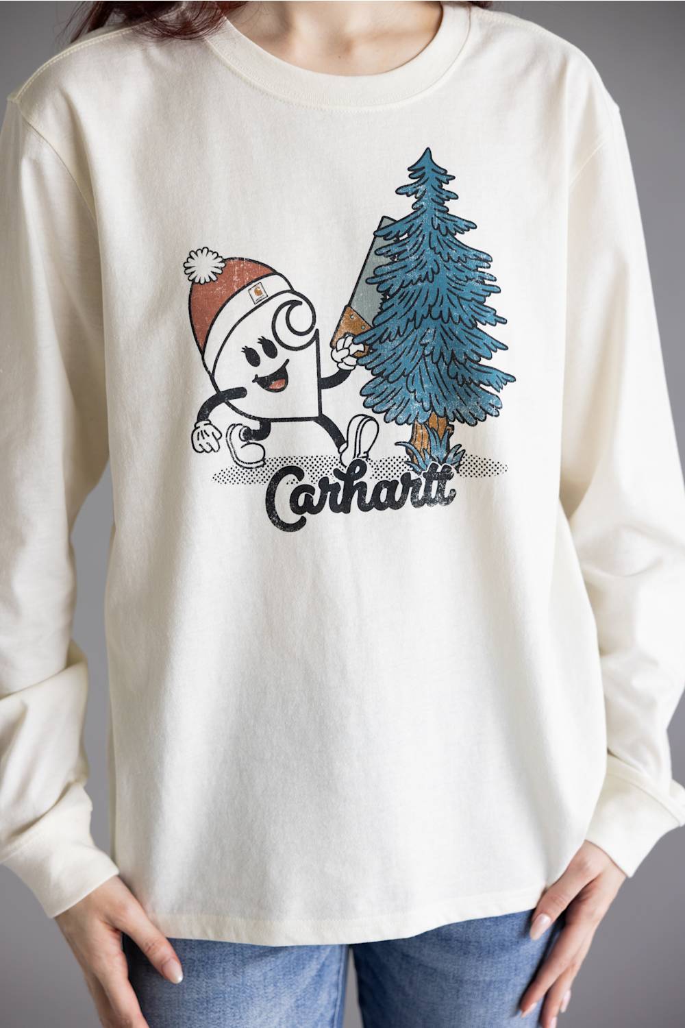A person wearing a cream-colored long-sleeve graphic t-shirt with a Carhartt logo and a cartoon character next to a pine tree, paired with blue jeans.