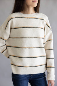 A woman wearing a cream-colored knit sweater with horizontal stripes in black and tan, paired with dark blue jeans, shown against a plain gray studio background.