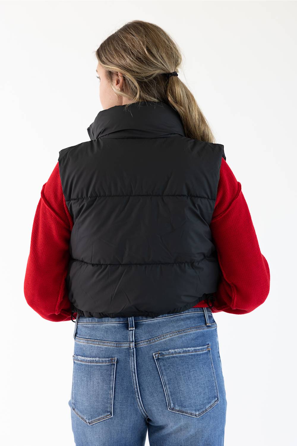 A woman viewed from behind wearing a black puffer vest over a red long-sleeve top and blue jeans, against a plain white studio background.